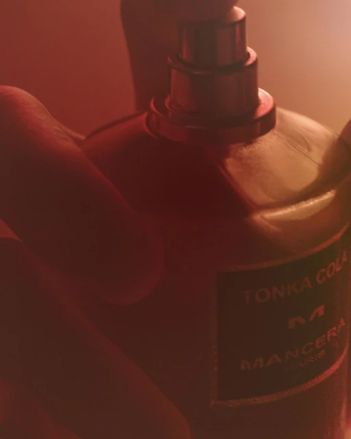 Close-up of a human hand holding a bottle of Tonka Cola Mancera perfume. The background is a blurred red.