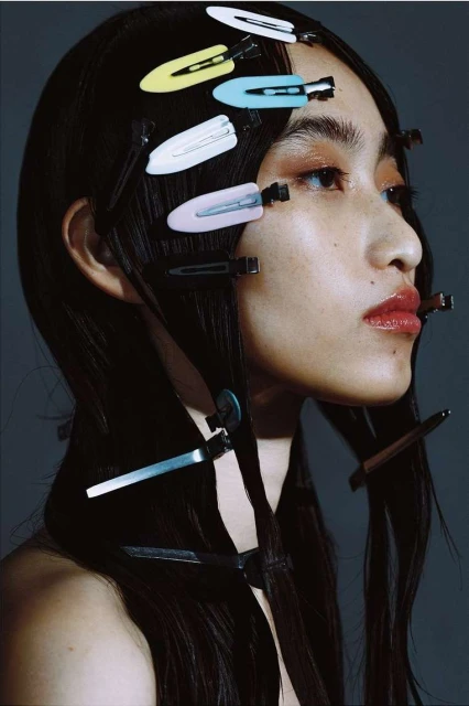 Close-up profile portrait of a young Asian woman with black hair styled with colorful hair clips, against a grey background in studio lighting.