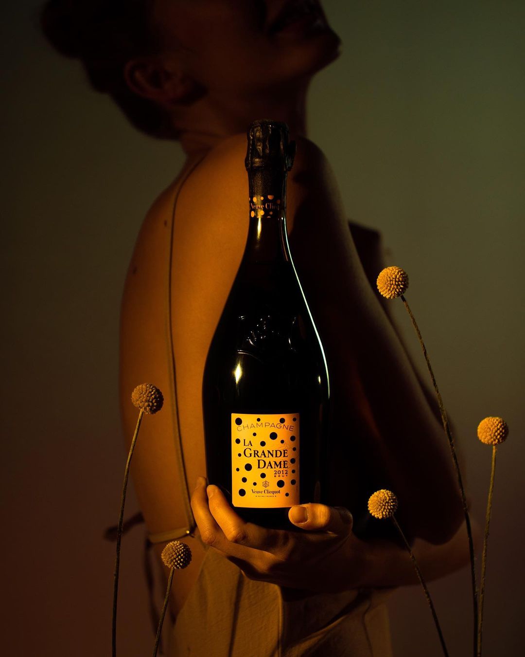 Champagne bottle held by a woman in dim lighting surrounded by small dried floral spheres on stems, against a grey background.