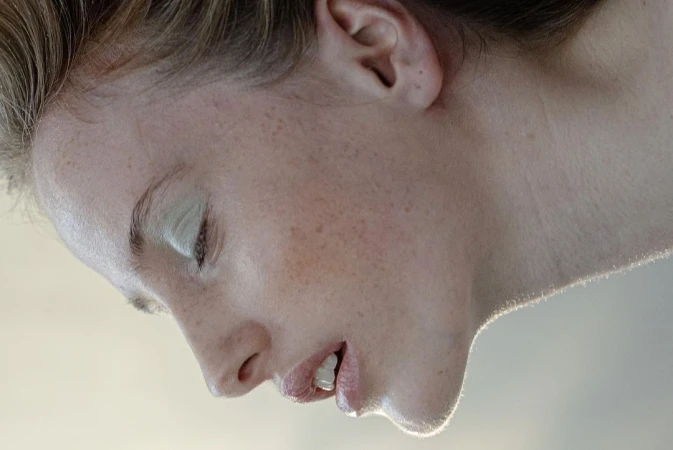 Close-up profile portrait of a woman with pale green eyeshadow, freckles, and parted lips against a neutral background.