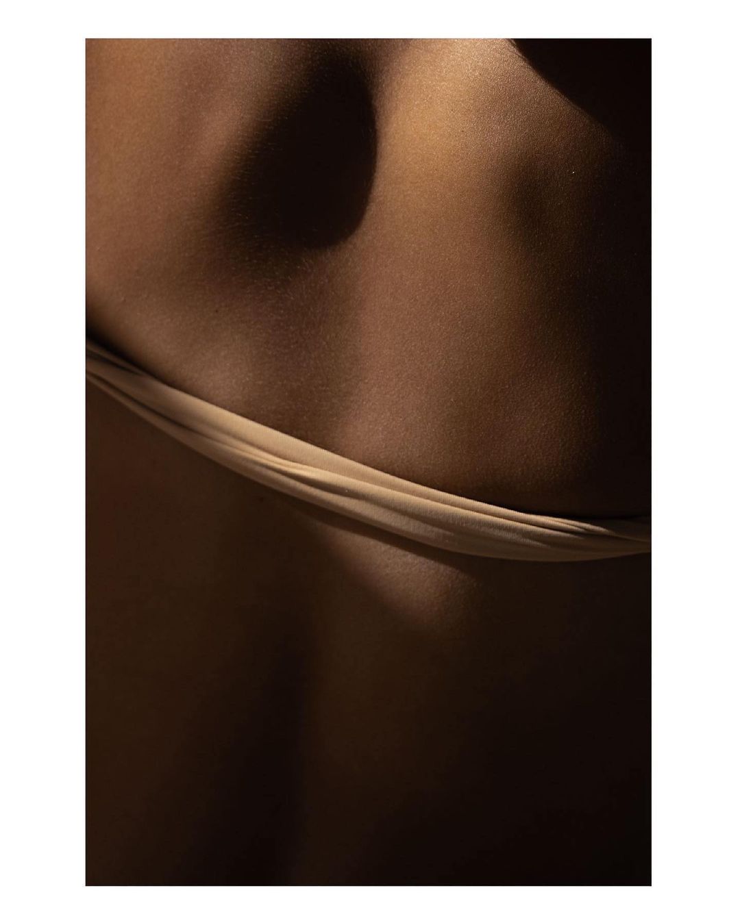 Close-up of the lower back of an adult female with a brown skin tone, wearing a beige fabric. The lighting is shadowy and dramatic.