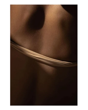 Close-up of the lower back of an adult female with a brown skin tone, wearing a beige fabric. The lighting is shadowy and dramatic.