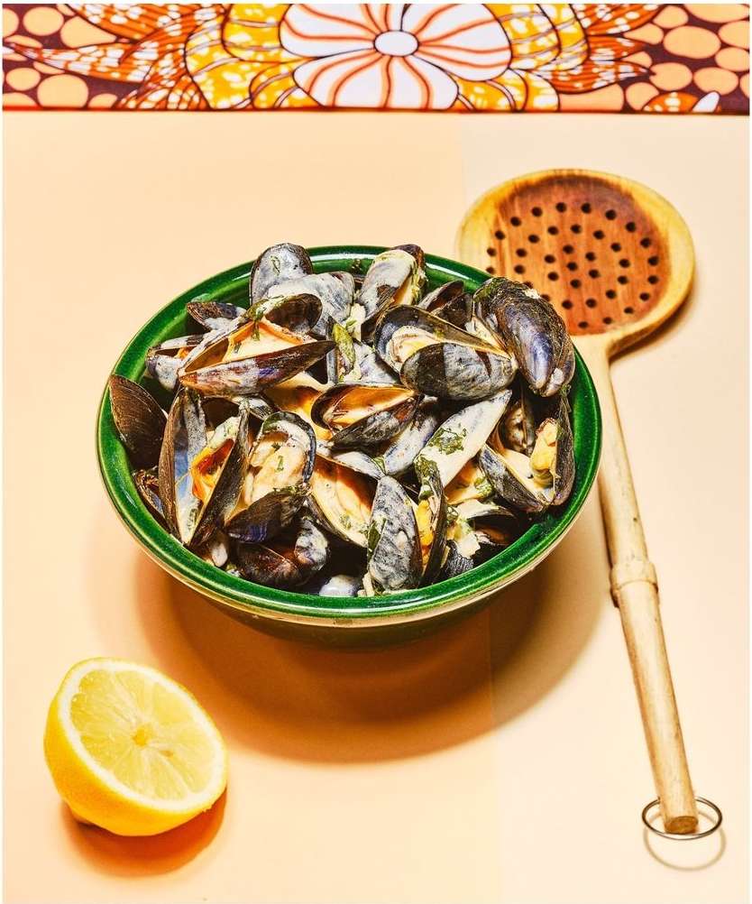 Still life photograph of a bowl of mussels in a creamy sauce next to a lemon half and wooden slotted spoon, on a peach surface.