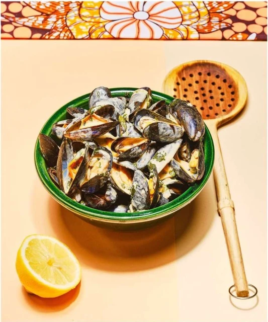 Still life photograph of a bowl of mussels in a creamy sauce next to a lemon half and wooden slotted spoon, on a peach surface.
