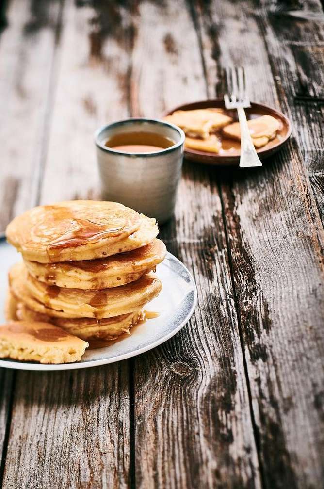 A stack of pancakes with syrup on a plate with a fork and a cup of tea, sitting on a rustic wooden table, food photography.