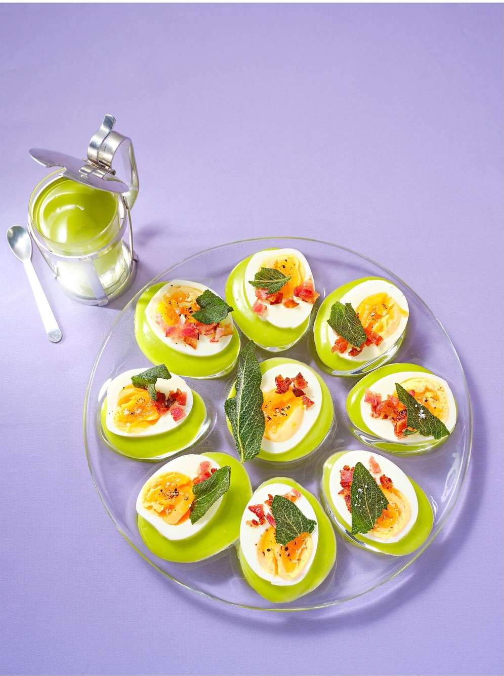 Deviled egg halves arranged on clear plate with sugar dispenser and spoon against light purple backdrop, food photography, close-up.