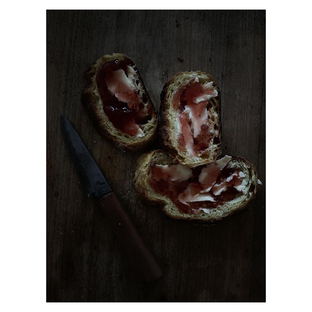 Overhead shot of three slices of bread topped with red jam and white spread, arranged next to a knife on a dark wooden surface.