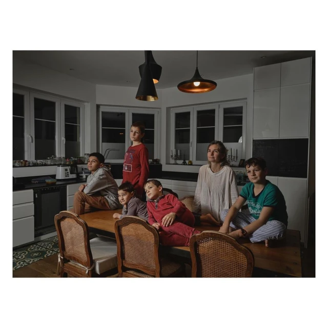 Group portrait in a kitchen of a woman and five children posing on a dining table with hanging lights, white cabinets, and windows.