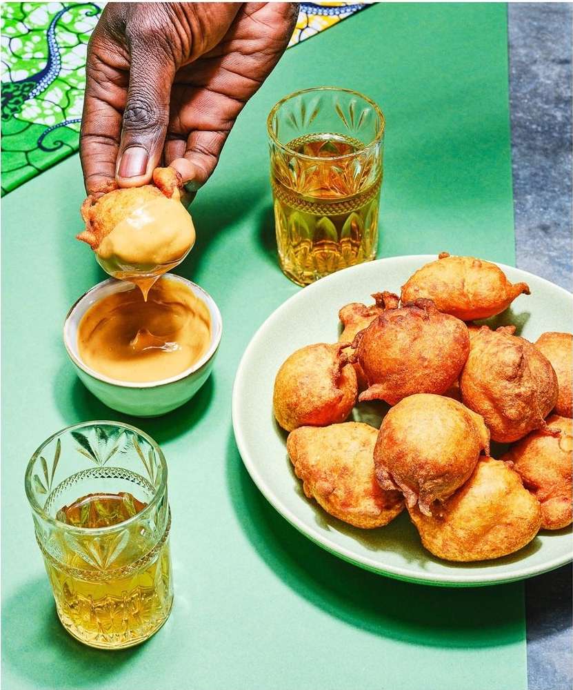 Person dips fried dough ball into sauce. Plate of dough balls, glasses of liquid, green tablecloth and background complete the food scene.