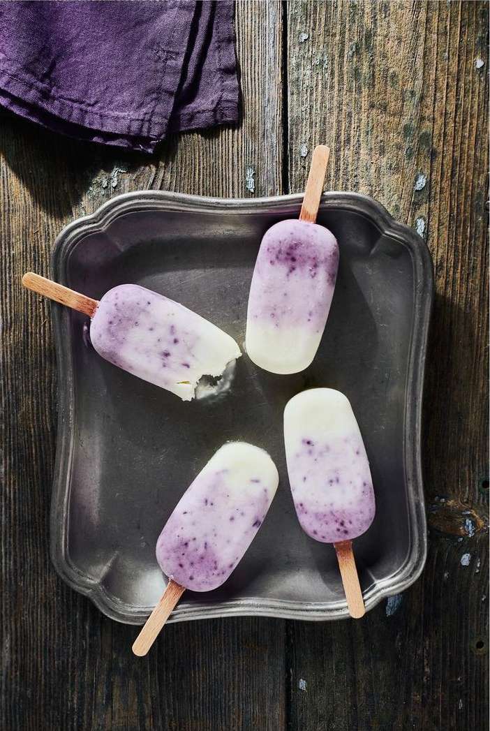 Overhead shot of four blueberry swirl ice popsicles on a silver platter atop a rustic wooden surface with a purple linen cloth.