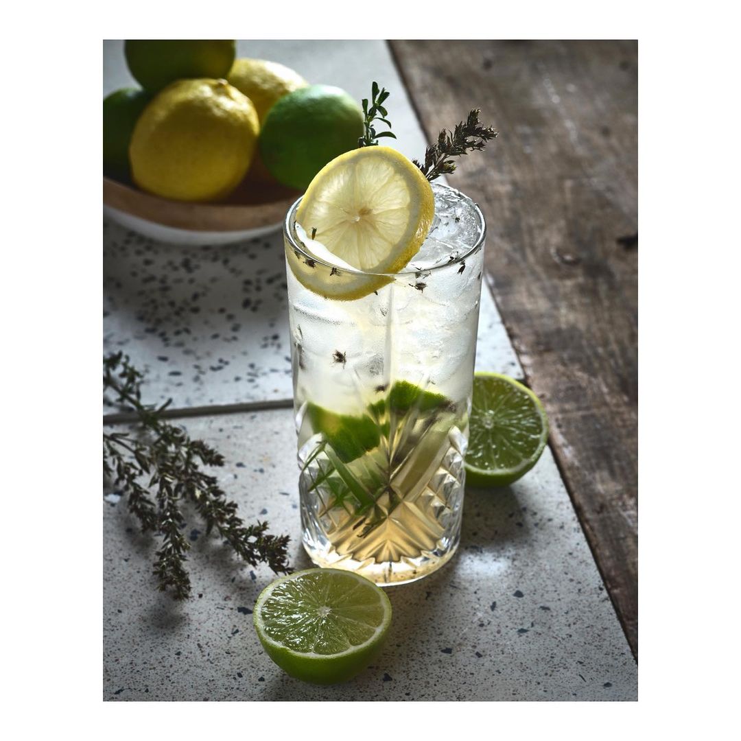 Refreshing citrus drink with ice in a textured glass, garnished with lemon slice and rosemary, alongside limes and lemons in a white bowl.