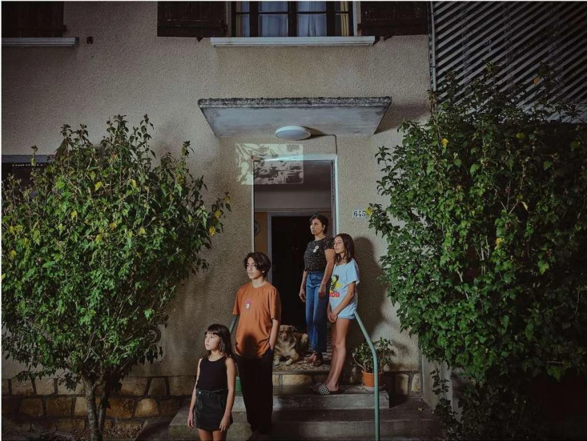 Four people and a dog stand on the porch of a house at night, illuminated by a porch light. Trees frame the scene.