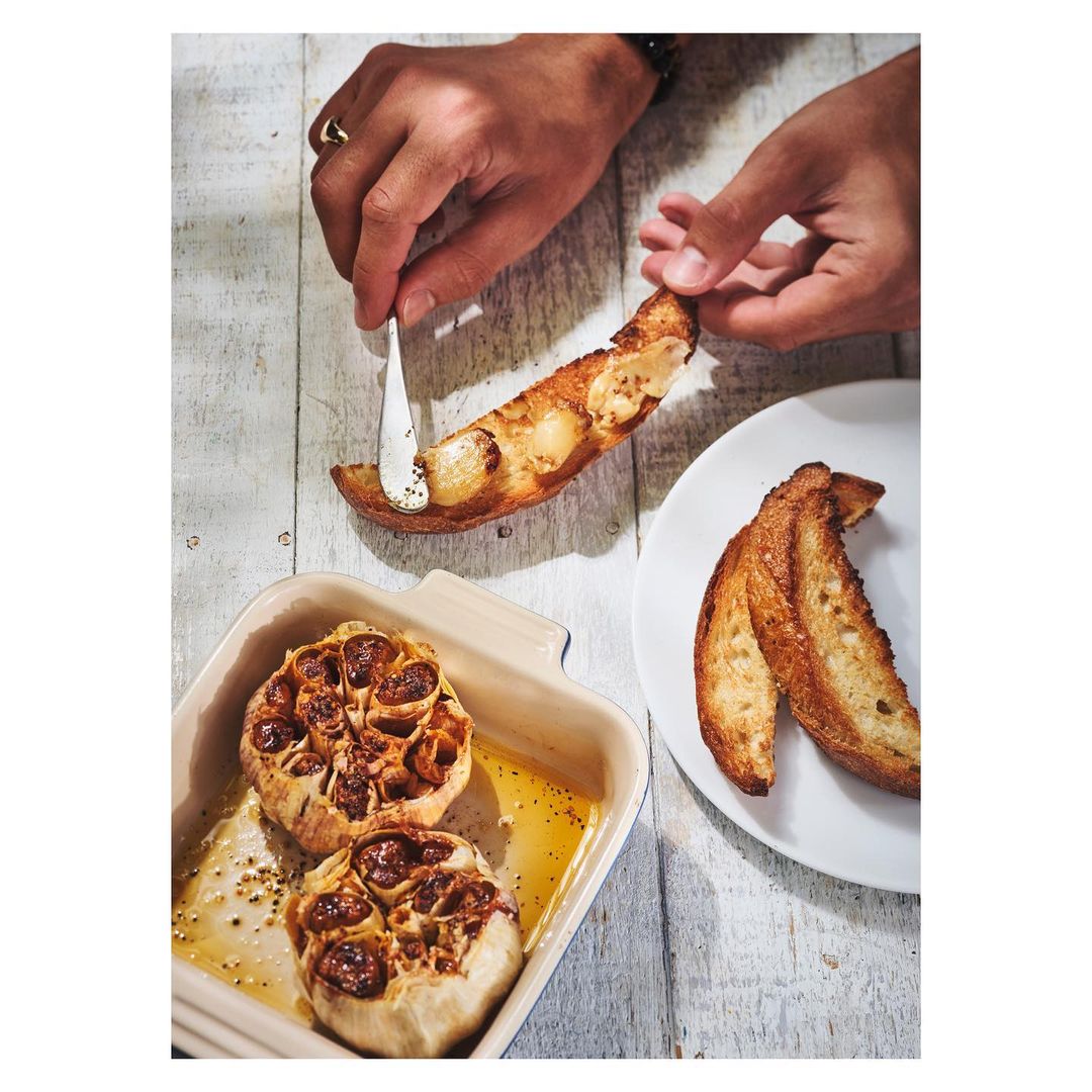 Person spreading garlic confit on toast. Roasted garlic bulbs in a ceramic dish sit beside bread slices on a white wooden table.