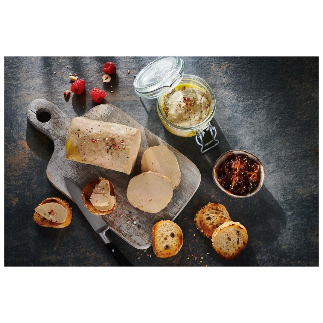 Overhead shot of foie gras, toast, and preserves arranged on a rustic wooden board with fresh raspberries and shadow patterns.