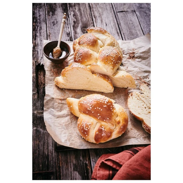 Overhead shot of challah bread with sesame seeds, cut on parchment paper with honey dipper and brown bowl, rustic wooden background.