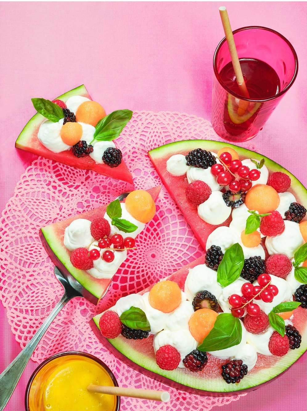 Overhead shot of watermelon 'pizza' slices decorated with whipped cream, fresh berries, cantaloupe, and basil, served on a pink backdrop.
