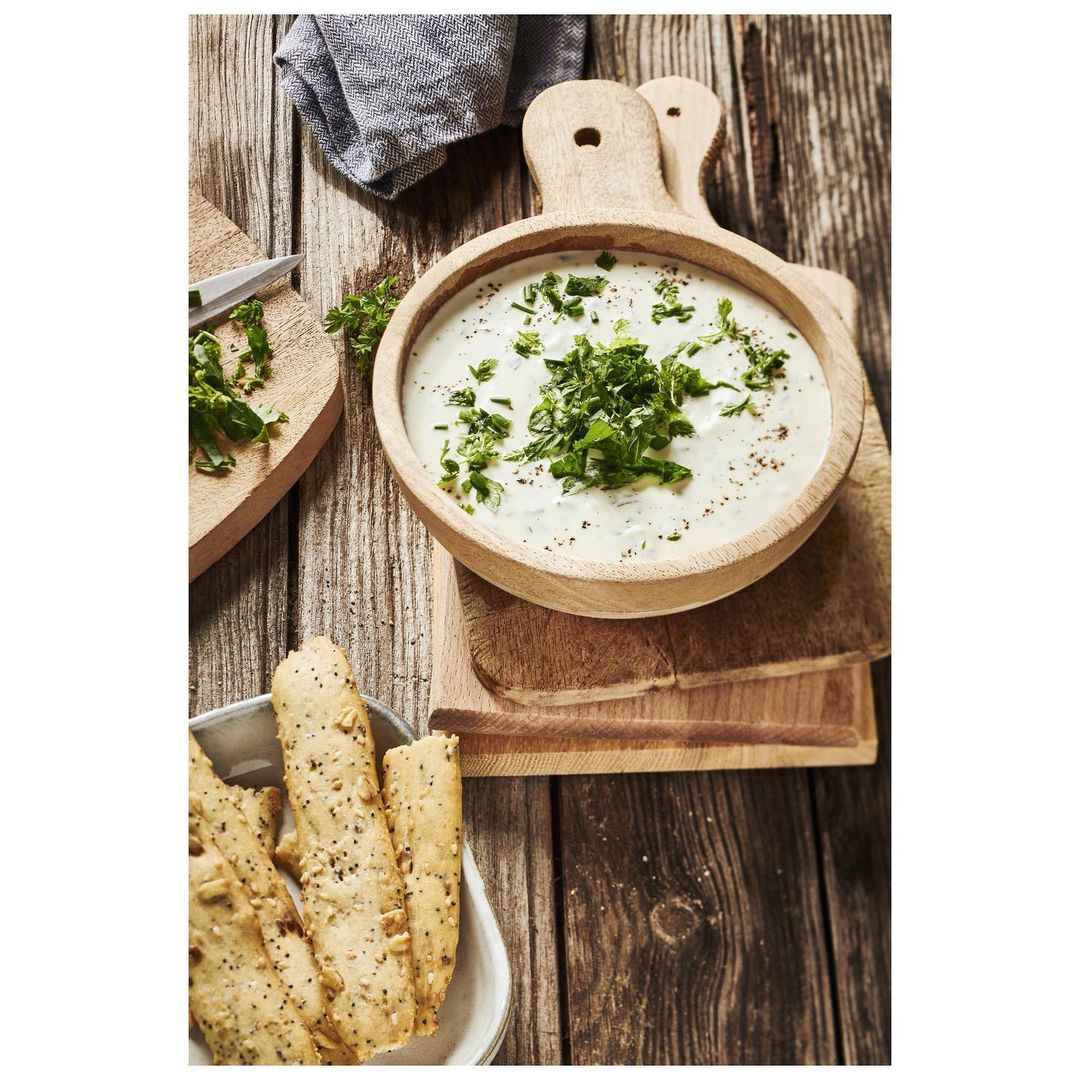 Overhead shot of a wooden bowl filled with creamy dip, sprinkled with herbs, sitting on a rustic wooden surface with breadsticks.