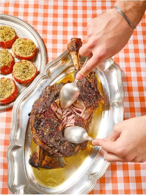 Overhead shot of a roasted lamb leg being carved on a silver platter next to stuffed tomatoes over a checkered tablecloth.