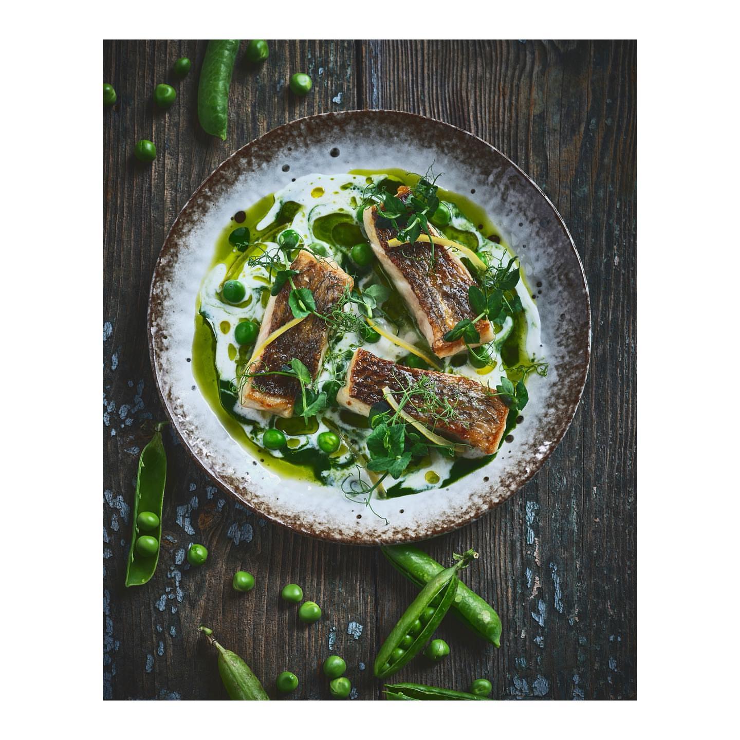 Overhead shot of a rustic plate with a fish dish garnished with fresh peas, green sauce, and herbs on a dark wooden surface.