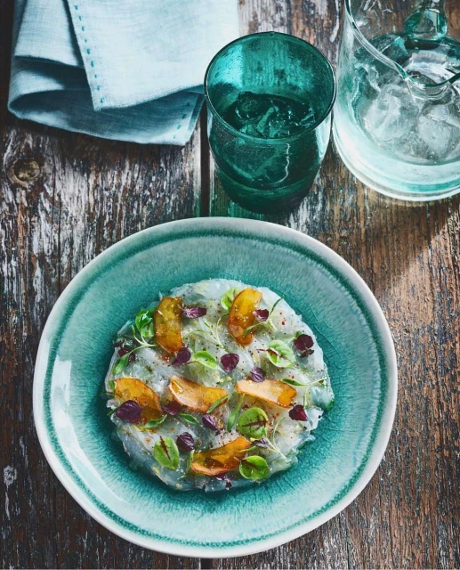 Overhead shot of fish carpaccio with fruit and herbs served on a teal plate, complemented by a linen napkin and a teal glass with ice.