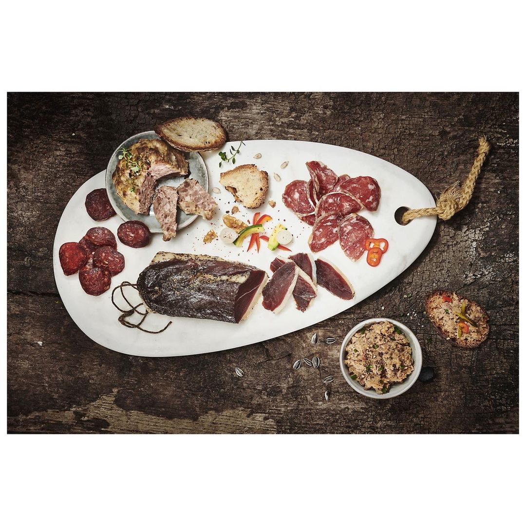 Overhead shot of an oval white platter with assorted meats, bread, and dips on a rustic wooden surface, creating a culinary presentation.