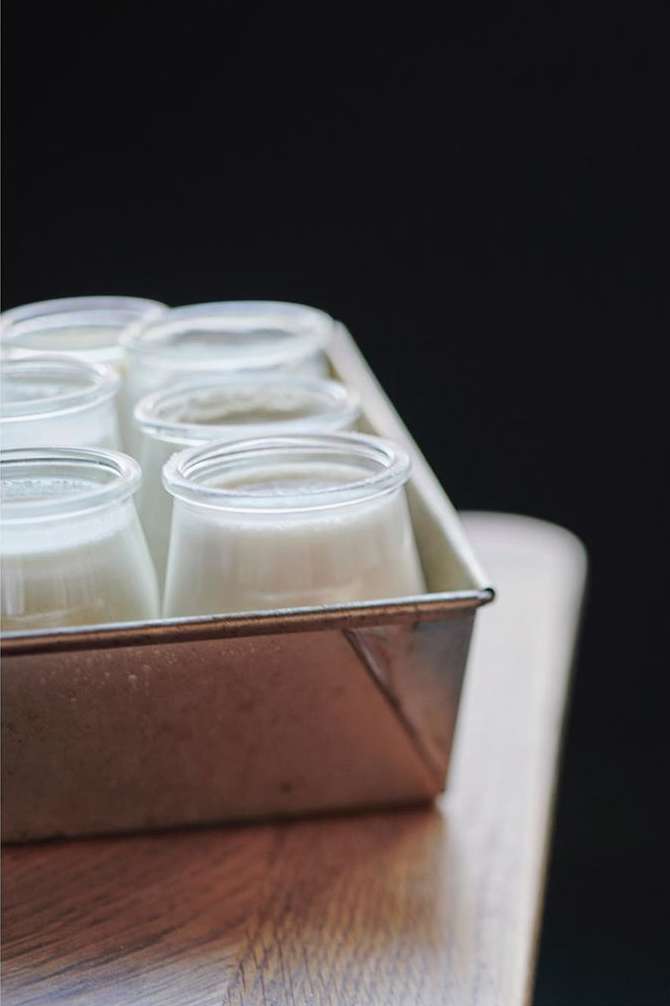 Close-up of glass jars filled with a creamy white substance in a metal baking tin, sitting on a wooden surface against a dark background.