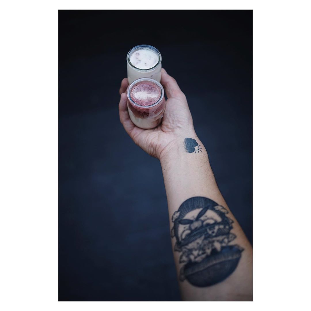Two glasses of smoothies held in an arm with tattoo art against a dark grey background. The smoothies are white and pink.