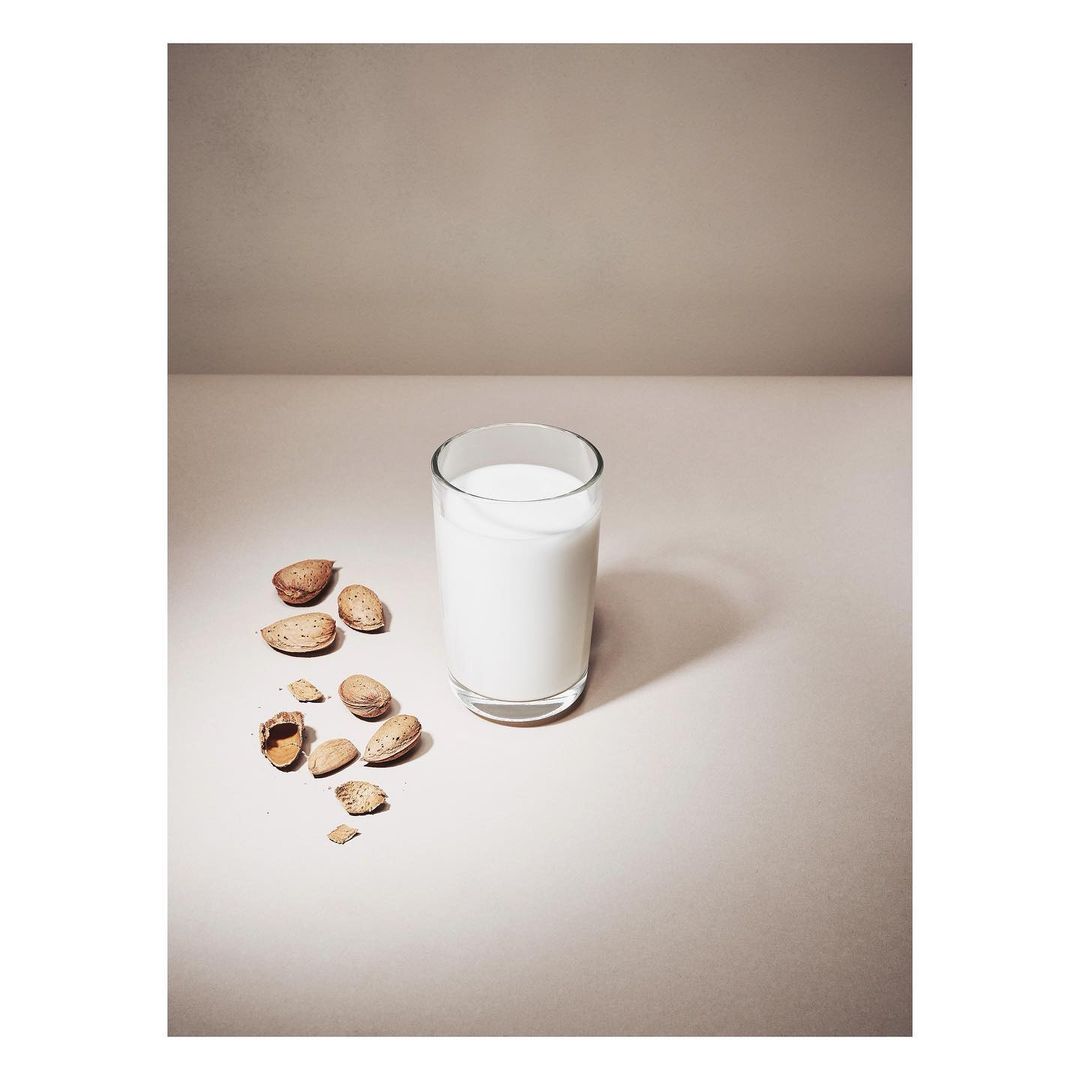 Almond milk in a clear glass sits beside shelled almonds on a neutral surface against a plain background, illuminated by soft lighting.