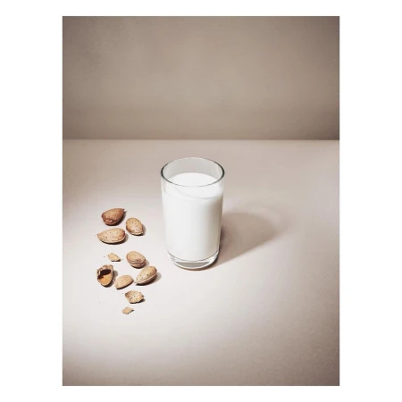 Almond milk in a clear glass sits beside shelled almonds on a neutral surface against a plain background, illuminated by soft lighting.