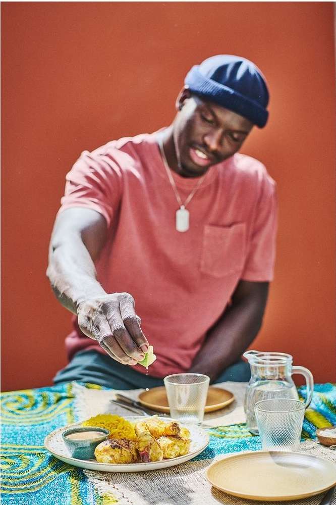 Dark-skinned man squeezes lime over chicken and rice dish at a table with plates and glasses. He wears a t-shirt and a dark-blue beanie.