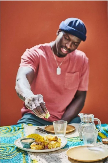 Dark-skinned man squeezes lime over chicken and rice dish at a table with plates and glasses. He wears a t-shirt and a dark-blue beanie.