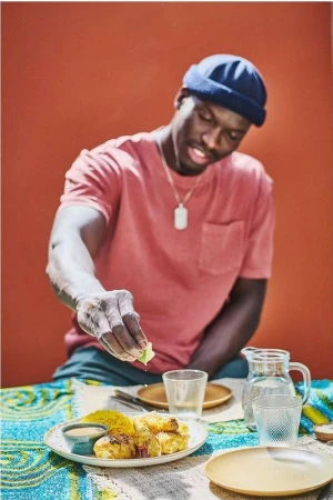 Dark-skinned man squeezes lime over chicken and rice dish at a table with plates and glasses. He wears a t-shirt and a dark-blue beanie.