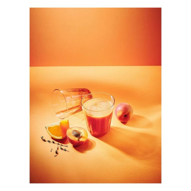 Still life of a full glass of juice, cut fruit (apricot and orange), and an empty glass against a bright orange background.