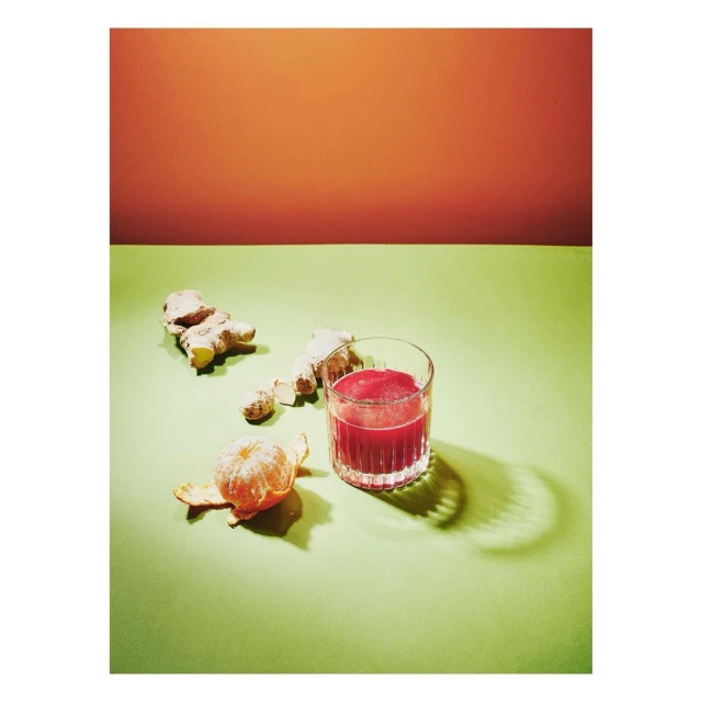 Still life shot of a glass of red juice, two pieces of ginger, and a peeled tangerine on a green table with an orange background.