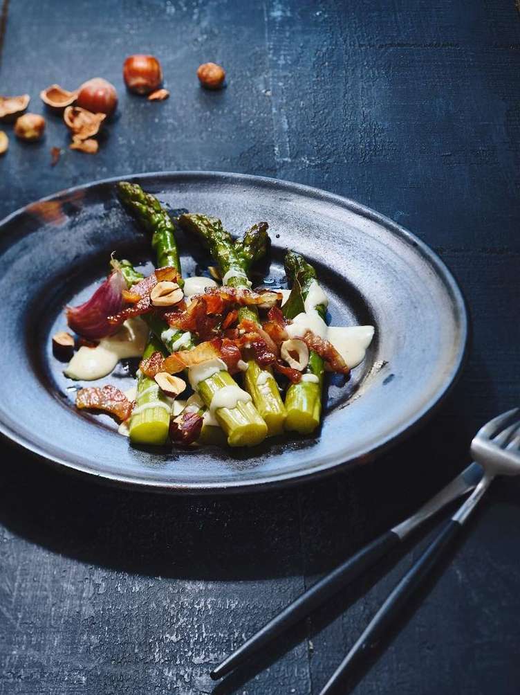 Asparagus spears on a dark plate with bacon and sauce, accompanied by a pair of black-handled forks on a dark surface.
