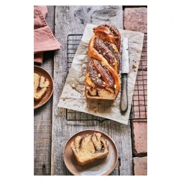 Overhead food photography of braided chocolate babka loaf on parchment paper with a sliced piece on a small ceramic plate.