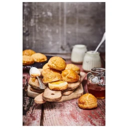 Freshly baked scones on wooden boards with a jar of jam, small spoons, and glass jars in a rustic setting.