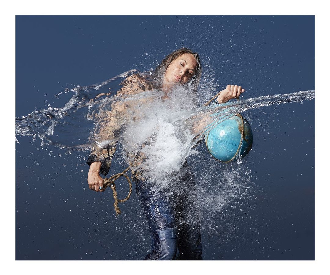 Woman in jeans, long sleeve top holding a blue and brown globe with a rope handle as water splashes on her against a blue background.
