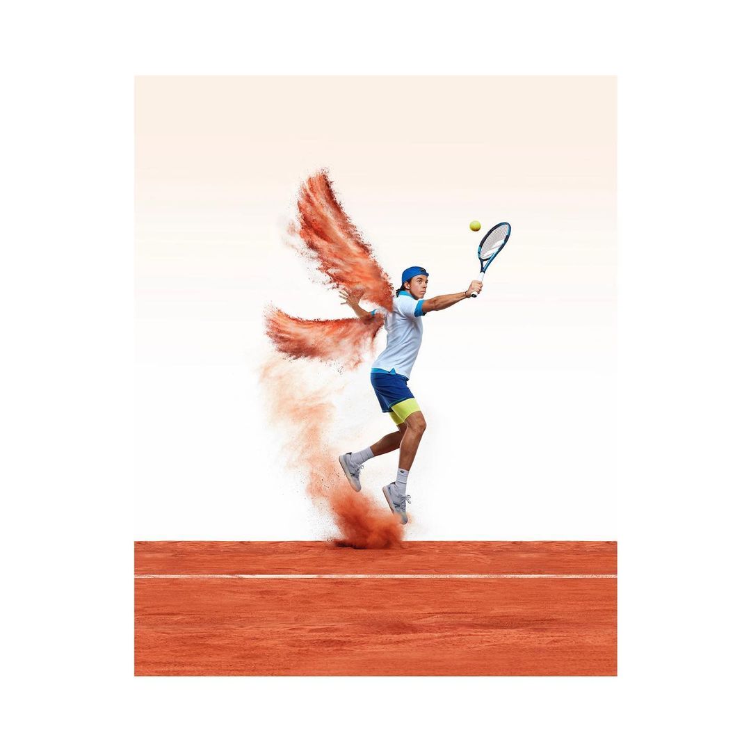 A tennis player hits a ball on a clay court while jumping in the air with an orange dust cloud in the shape of wings behind him.