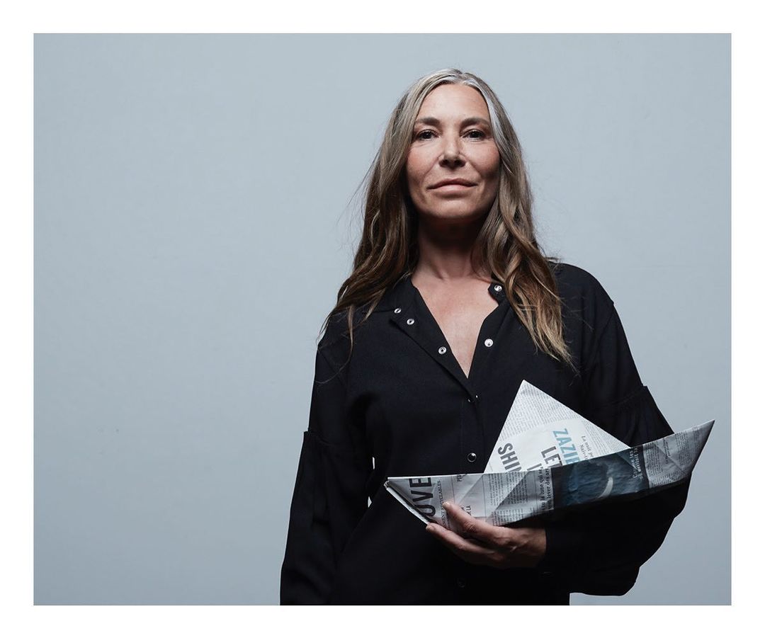 Portrait of a mature woman holding a paper boat against a grey background in a studio, wearing a black shirt and with long grey hair.