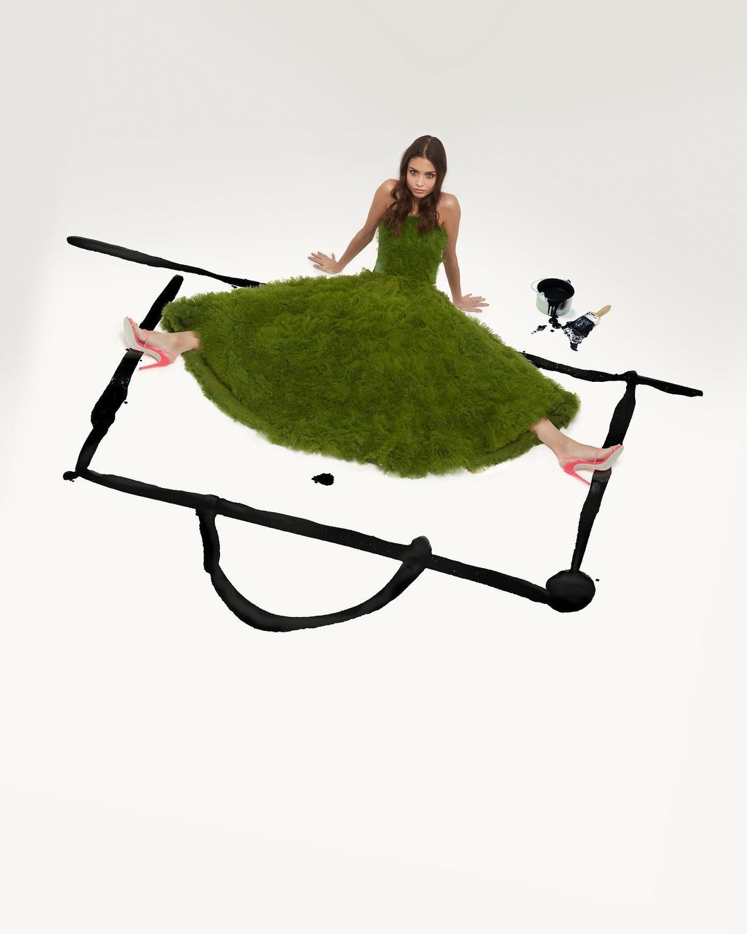 Woman in green grass dress posing on white backdrop with black paint, wearing pink high heels. Fashion editorial shot with artist style.