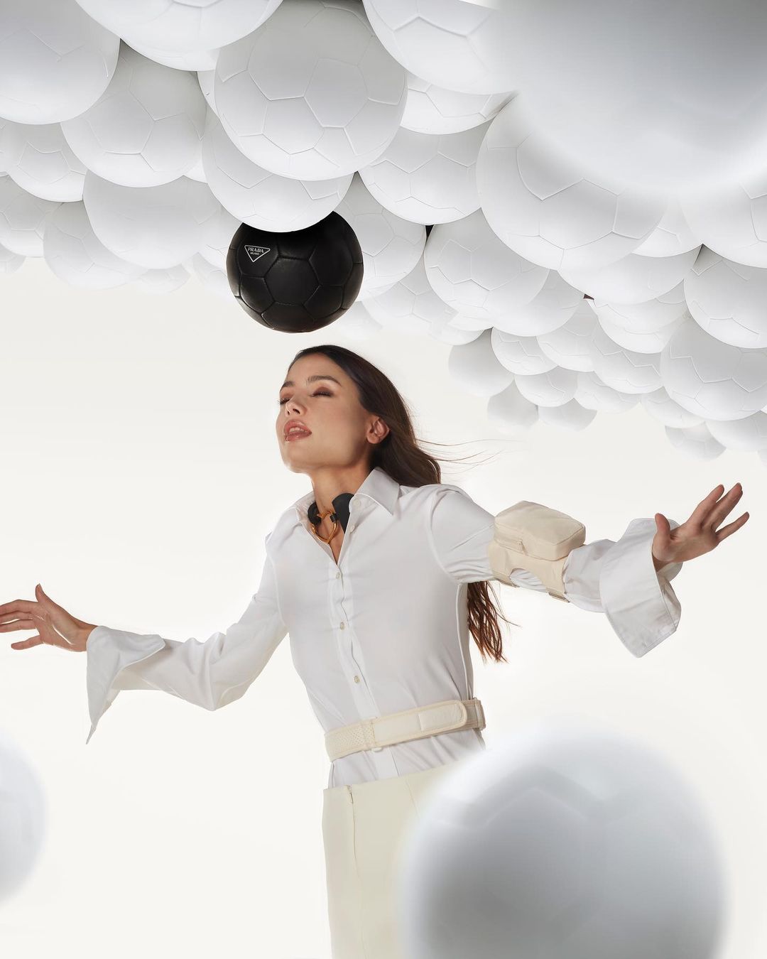 Woman in white outfit poses with arms outstretched among white and one black soccer ball. Low angle, fashion editorial.
