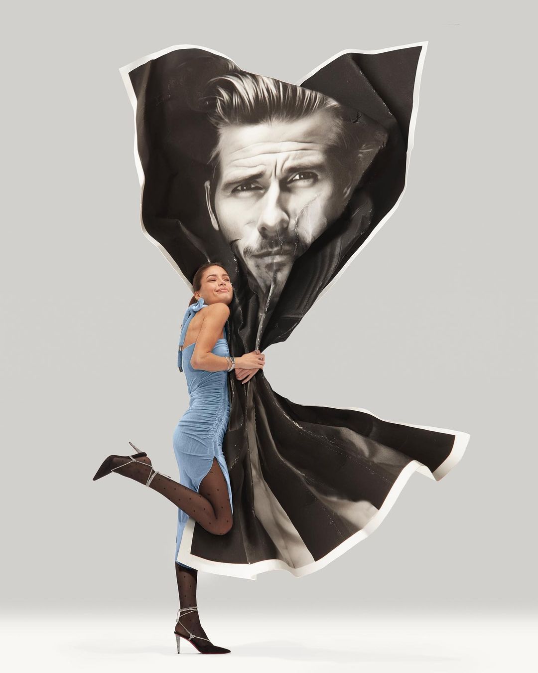 Woman in blue dress poses playfully holding a large black and white portrait against a pale grey background in a studio shot.
