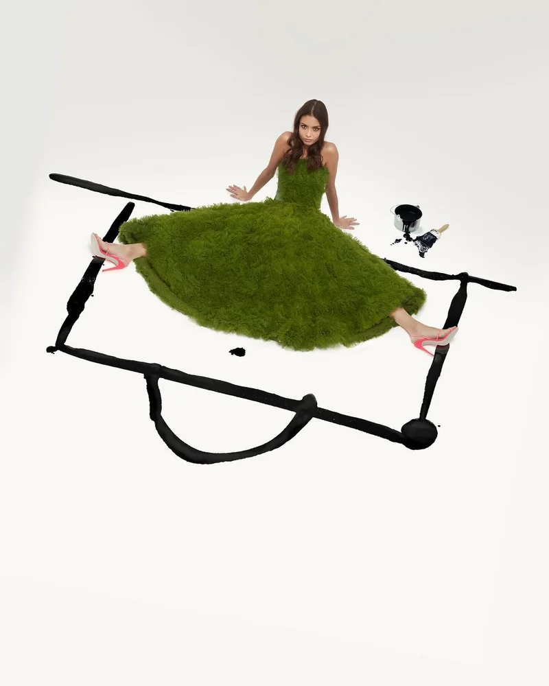 Model in green textured grass dress wearing pink high heels seated inside a painted black frame on white floor