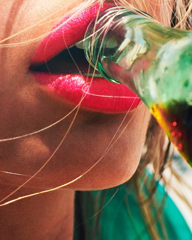 Close-up of a woman's face as she drinks from a green glass bottle, her red lipstick contrasting with the bright bottle.