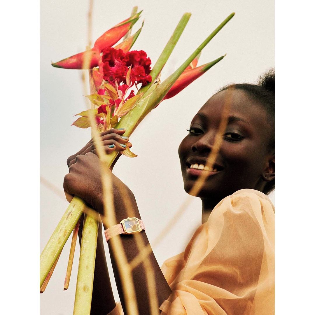 Portrait of a smiling Black woman holding flowers with a gold watch, framed by out-of-focus stems. Light peach dress, neutral backdrop.