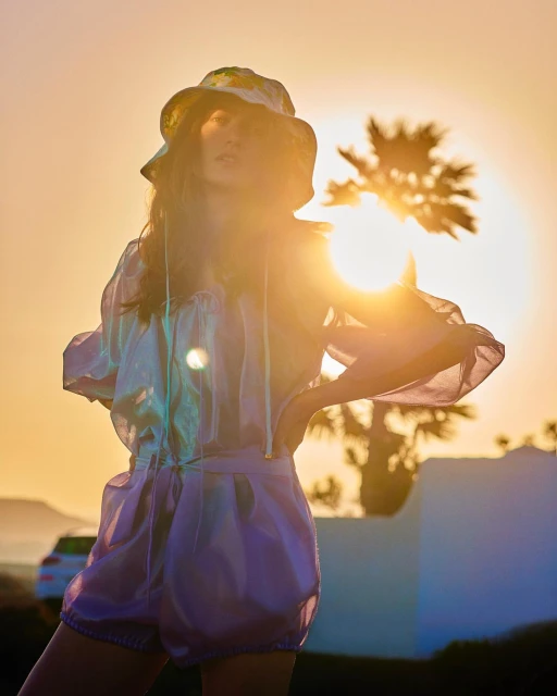 Young woman in a bucket hat and sheer top, posing in golden hour sunlight with palm trees and a car in the background.