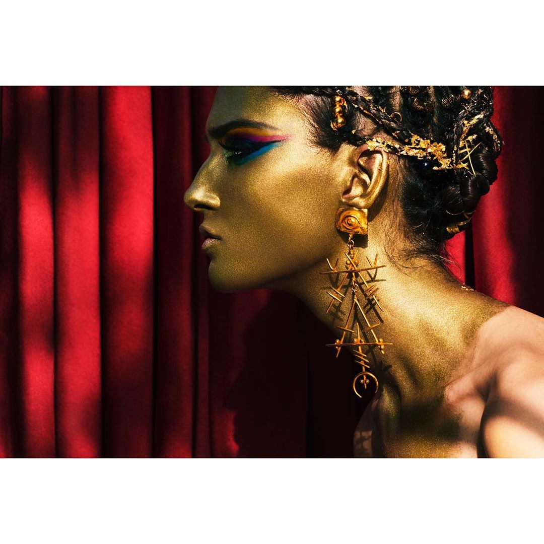 Profile of a model with gold makeup wearing ornate earrings against a red curtain backdrop, showcasing beauty and fashion.