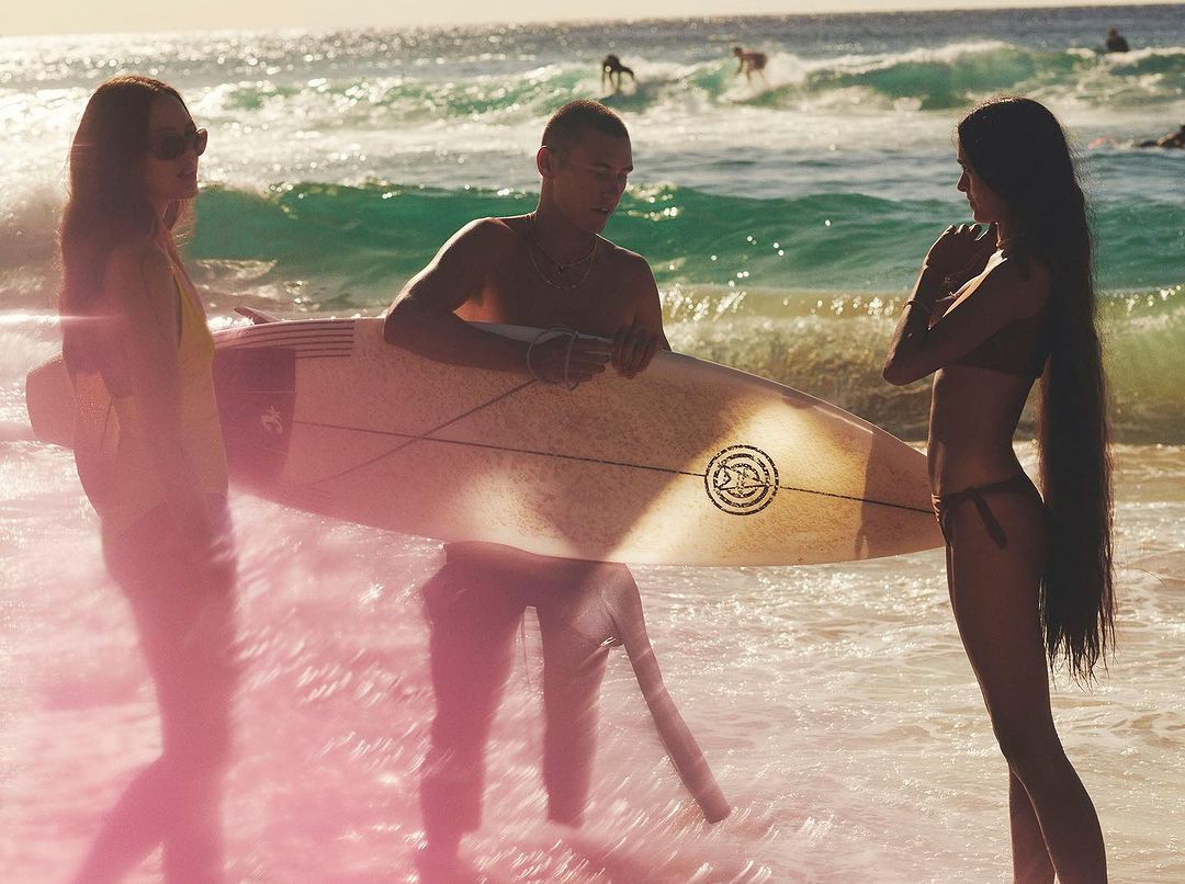 Three people standing on a beach near ocean waves, one holding a surfboard, with other surfers in the background on the water.