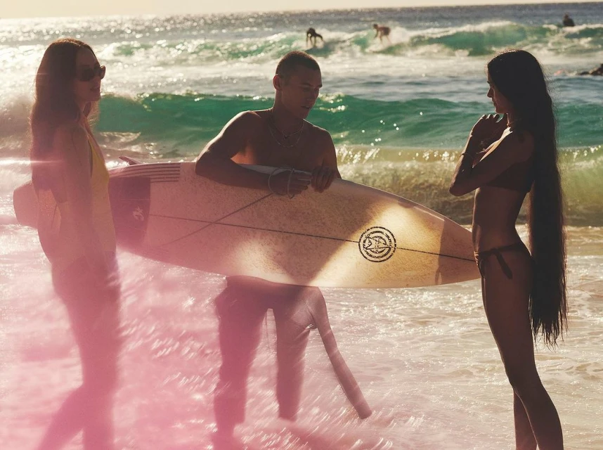 Three people standing on a beach near ocean waves, one holding a surfboard, with other surfers in the background on the water.
